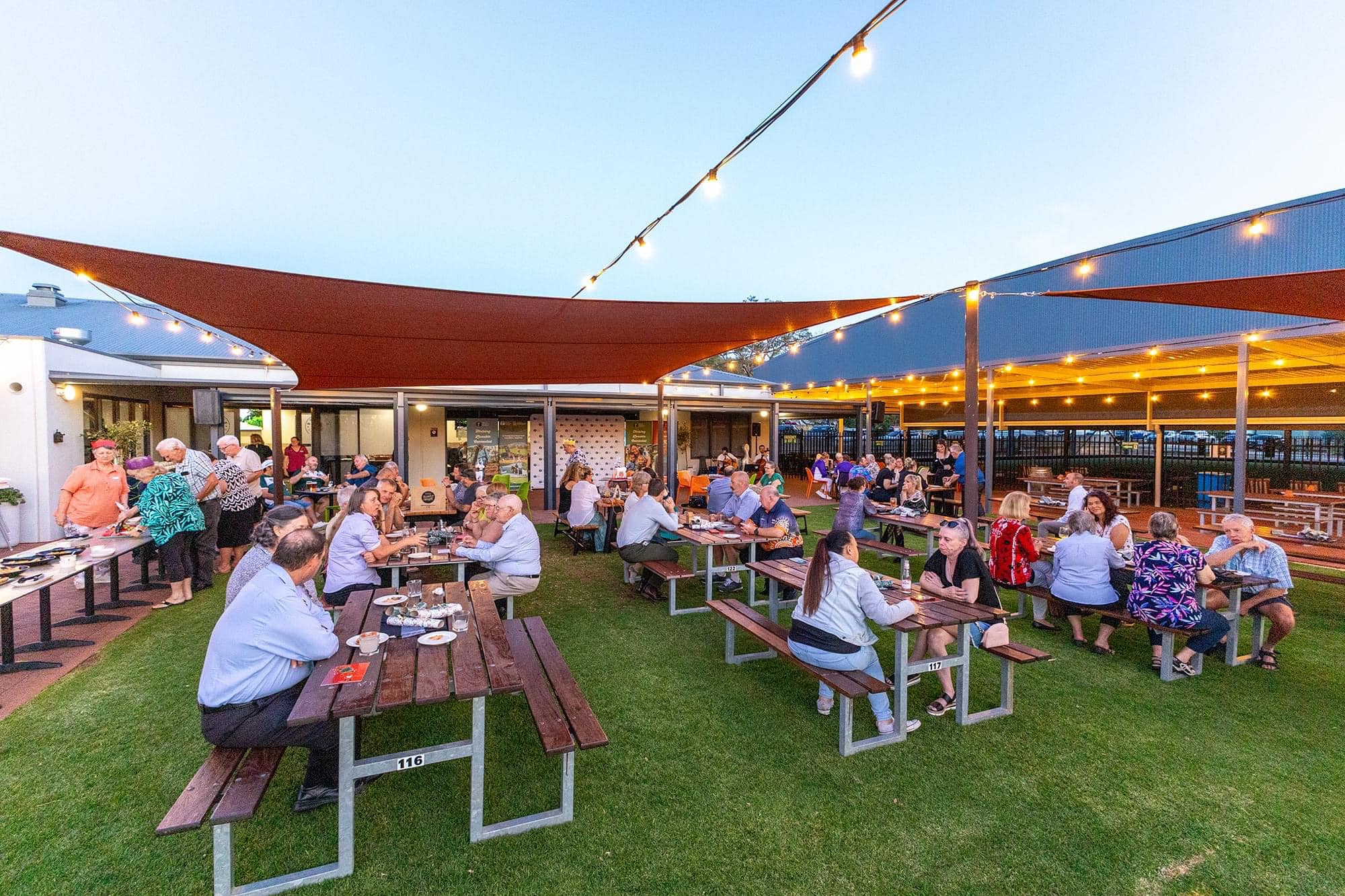 Outdoor dining area with guests seated at picnic tables under lights
